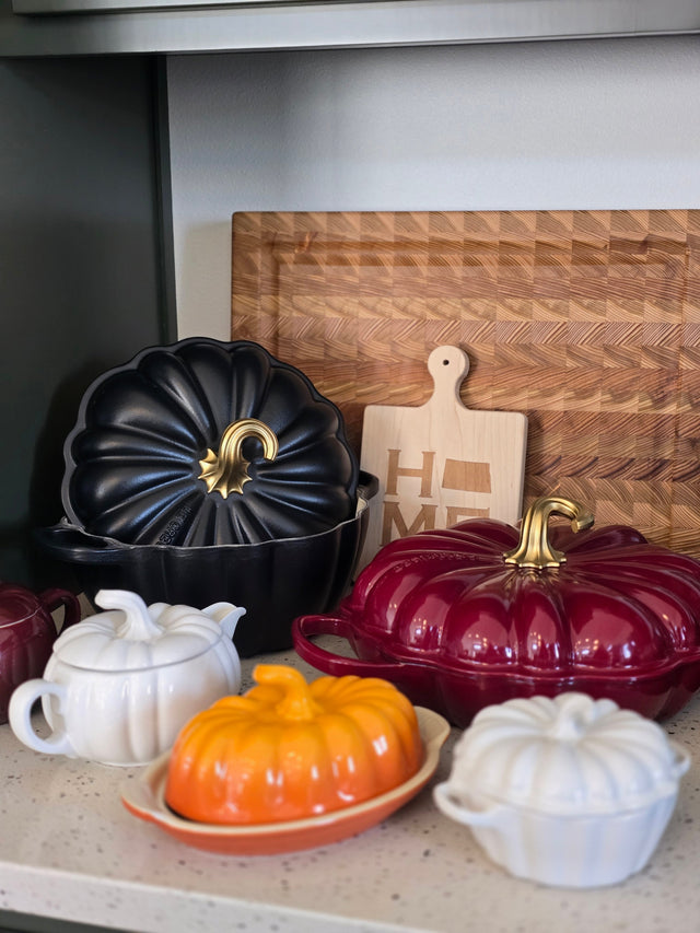 a collection of kitchen utensils and containers, including a wooden cutting board, a black pot lid, a red pot lid, a white teapot, and a wooden tray.