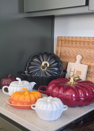 a collection of colorful, decorative kitchenware, including a red and black pot, a black pot with a gold handle, and several white and orange pots, all arranged on a kitchen counter.