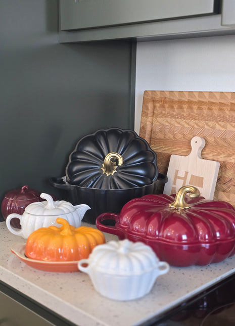 a collection of colorful, decorative kitchenware, including a red and black pot, a black pot with a gold handle, and several white and orange pots, all arranged on a kitchen counter.