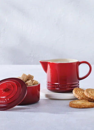 a red ceramic pitcher, a red ceramic container with a lid, and a plate of cookies on a white surface.